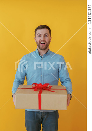 Surprised, holding gift box. Attractive man in blue shirt is against yellow background 131381385