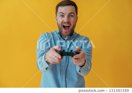 Playing video game with joystick. Attractive man in blue shirt is against yellow background Playing video game with joystick. Attractive man in blue shirt is against yellow background 131381389