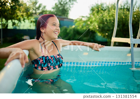 Teen girl enjoying a summer afternoon in a backyard pool surrounded by lush greenery and sunshine Teen girl enjoying a summer afternoon in a backyard pool surrounded by lush greenery and sunshine 131381554