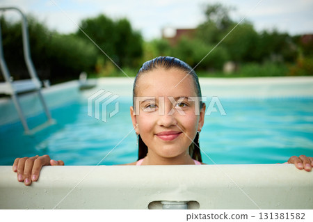 Teen girl enjoys summer fun while peeking over the edge of a swimming pool in a sunny backyard setting Teen girl enjoys summer fun while peeking over the edge of a swimming pool in a sunny backyard setting 131381582