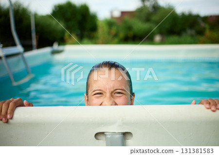 Teen girl enjoys summer fun while peeking over the edge of a swimming pool in a sunny backyard setting Teen girl enjoys summer fun while peeking over the edge of a swimming pool in a sunny backyard setting 131381583