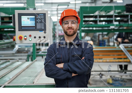 Beautiful man is standing with arms crossed. Factory worker is indoors with hard hat Beautiful man is standing with arms crossed. Factory worker is indoors with hard hat 131381705