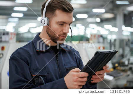 In headphones, with tablet. Factory worker is indoors with hard hat In headphones, with tablet. Factory worker is indoors with hard hat 131381786