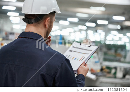 Back view, with notepad and graphs. Factory worker is indoors with hard hat Back view, with notepad and graphs. Factory worker is indoors with hard hat 131381804