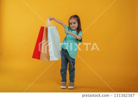 Shopping bags in hand. Cute little girl is against yellow background 131382387