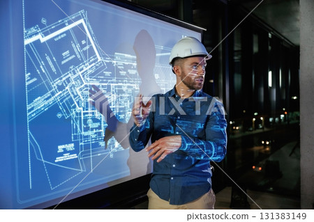 Protective hard hat on head. Man architect in hard hat is near projector with construction plan 131383149