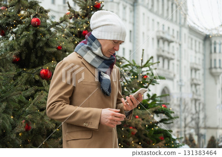 Adult handsome man stands thoughtfully with phone on street near Christmas trees in winter attire in modern city. Xmas holiday. 131383464