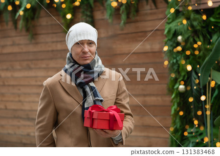 Winter portrait of caucasian man wearing beige coat stand with Christmas red gift box at modern city. Cold season. Outdoors. Xmas holiday. Winter portrait of caucasian man wearing beige coat stand with Christmas red gift box at modern city. Cold season. Outdoors. Xmas holiday. 131383468
