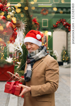Adult happy man in Santa hat buying red gift at Christmas outdoor market. Xmas shopping. Side view. Holiday spirit. Close up. 131383469