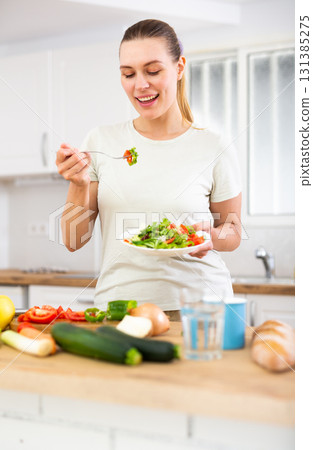 Vegeterian woman standing at table in kitchen and eating vegetable salad 131385275