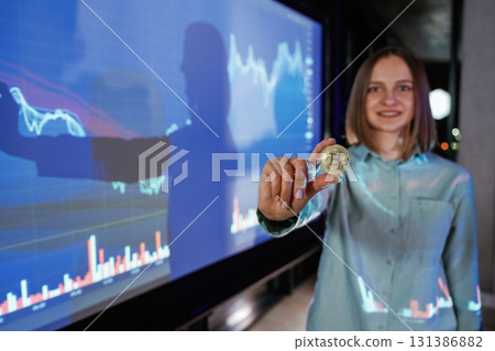 Close up of digital currency coin in hand. Businesswoman is against projector with graphs, indoors Close up of digital currency coin in hand. Businesswoman is against projector with graphs, indoors 131386882