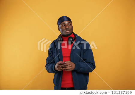 Smartphone in hands, headphones on the neck. Handsome black man is in the studio against yellow background 131387199