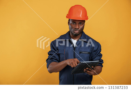 Digital tablet in hand, construction worker. Handsome black man is in the studio against yellow background Digital tablet in hand, construction worker. Handsome black man is in the studio against yellow background 131387233