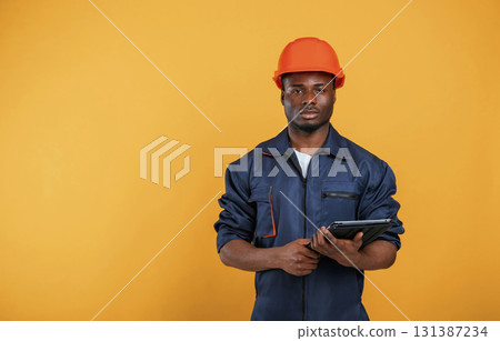 Digital tablet in hand, construction worker. Handsome black man is in the studio against yellow background 131387234