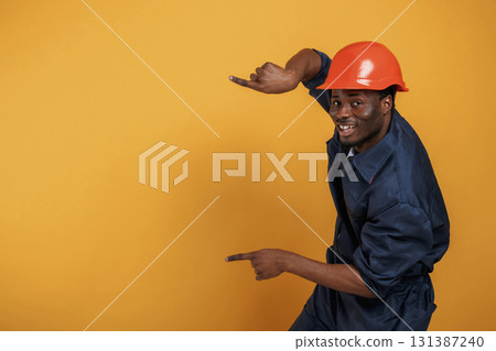 Guy in hard hat showing copy space by fingers. Handsome black man is in the studio against yellow background 131387240