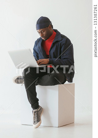 Sitting and using laptop. Black man in cap is in the studio against white background 131387261