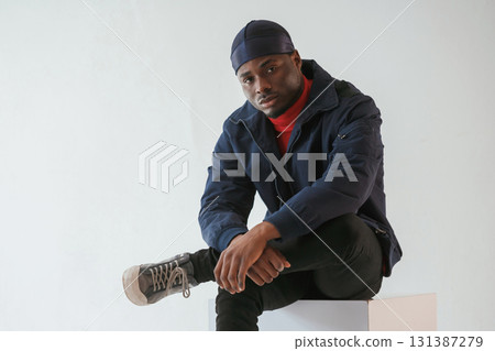 Sitting on the cube chair. Black man in cap is in the studio against white background 131387279