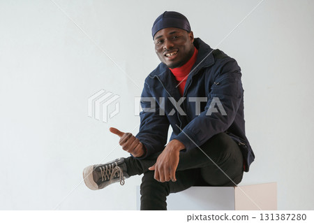 Calm, sitting, smiling and showing thumb up. Black man in cap is in the studio against white background 131387280