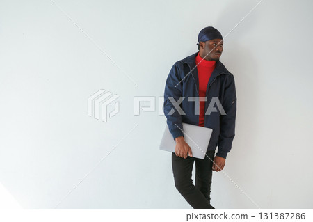 Standing and holding laptop in hand. Black man in cap is in the studio against white background 131387286