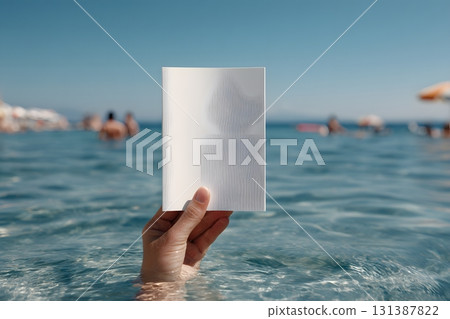 A hand holds a blank white card above blue water with people in the background. 131387822