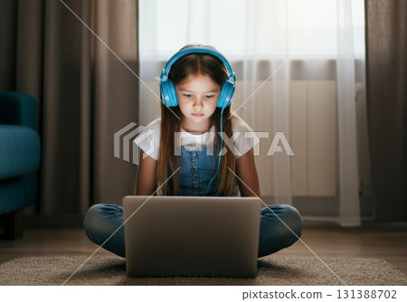 Young girl with headphones focused on laptop in cozy living room with soft lighting 131388702