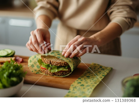 Hands wrapping sandwich in reusable beeswax wrap on wooden cutting board in kitchen setting 131389368
