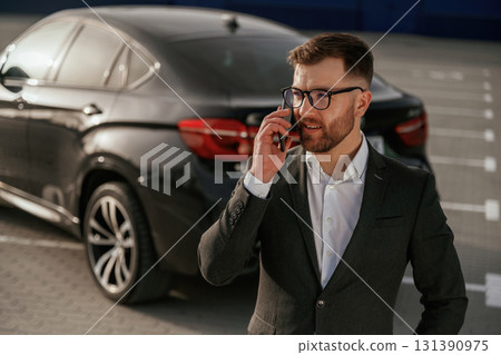 Talking by using phone. Businessman in suit is near his black car outdoors 131390975