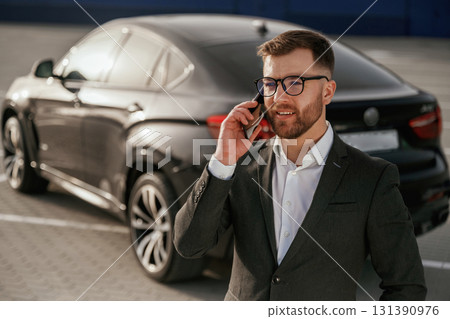 Talking by using phone. Businessman in suit is near his black car outdoors Talking by using phone. Businessman in suit is near his black car outdoors 131390976