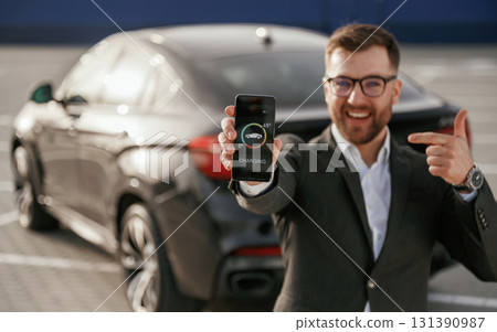Phone app that is monitoring charging. Businessman in suit is near his black car outdoors 131390987