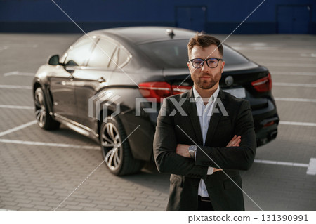 Standing with arms crossed. Businessman in suit is near his black car outdoors 131390991