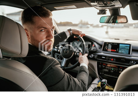 Looking behind at the camera. Man in black suit is sitting in the car, driving 131391013
