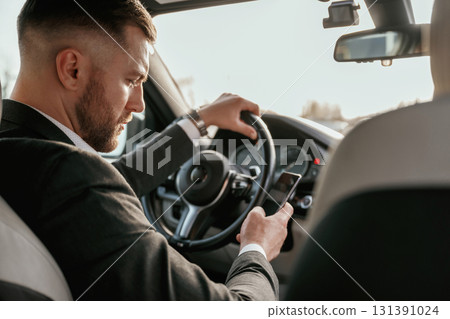 Surfing the internet by using smartphone. Man in black suit is sitting in the car, driving 131391024