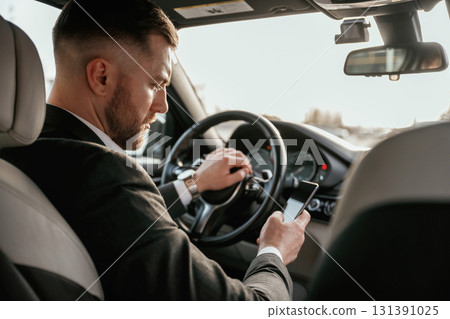 Surfing the internet by using smartphone. Man in black suit is sitting in the car, driving 131391025
