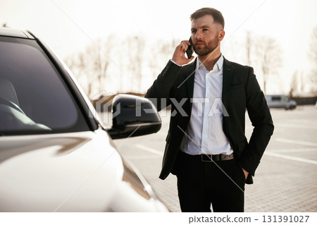 Talking by using phone. Businessman in suit is near his black car outdoors Talking by using phone. Businessman in suit is near his black car outdoors 131391027
