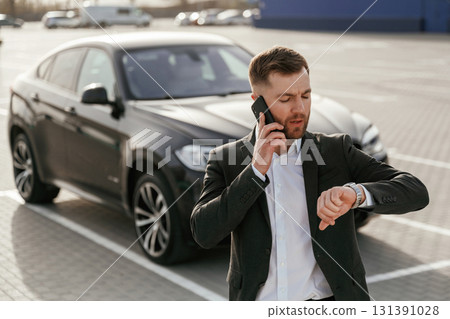 Talking by using phone. Businessman in suit is near his black car outdoors 131391028