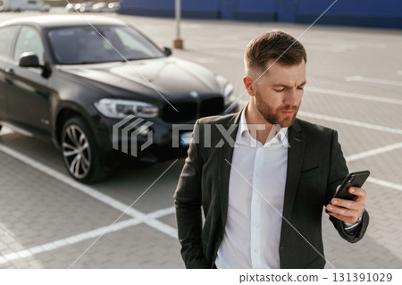 Smartphone in hand. Businessman in suit is near his black car outdoors 131391029