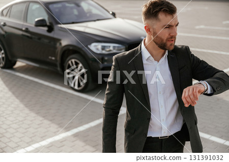 Checks the time. Businessman in suit is near his black car outdoors 131391032