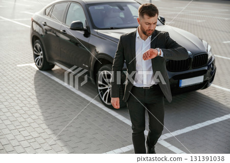Checks the time. Businessman in suit is near his black car outdoors Checks the time. Businessman in suit is near his black car outdoors 131391038