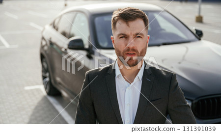 Focused portrait, against vehicle. Businessman in suit is near his black car outdoors Focused portrait, against vehicle. Businessman in suit is near his black car outdoors 131391039