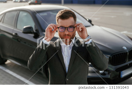 In glasses, front view. Businessman in suit is near his black car outdoors In glasses, front view. Businessman in suit is near his black car outdoors 131391043