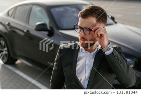 In glasses, front view. Businessman in suit is near his black car outdoors In glasses, front view. Businessman in suit is near his black car outdoors 131391044