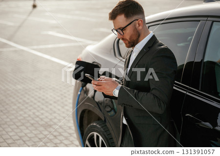 Leaning on the vehicle while it charges, with smartphone. Businessman in suit is near his black car outdoors 131391057
