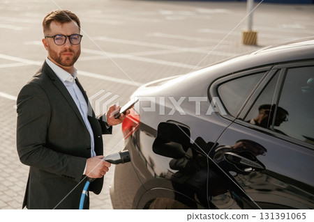 Holding wire for charging vehicle. Businessman in suit is near his black car outdoors 131391065