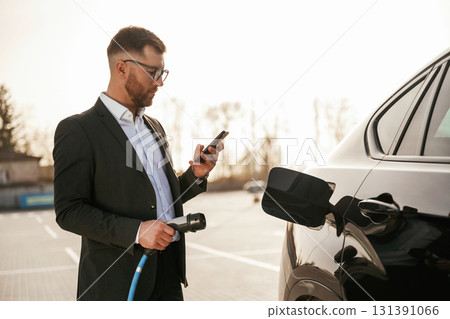 Holding wire for charging vehicle. Businessman in suit is near his black car outdoors 131391066
