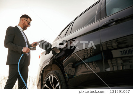 Charging the electric vehicle. Businessman in suit is near his black car outdoors 131391067