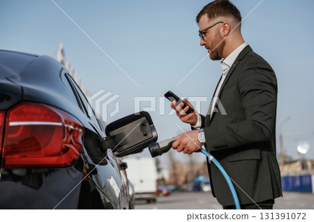 Natural lighting, day. Businessman in suit is near his black car outdoors 131391072