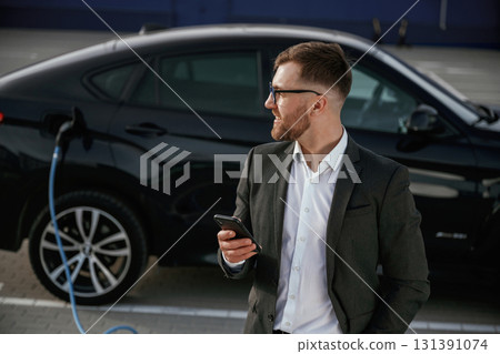 Smartphone in hand. Businessman in suit is near his black car outdoors Smartphone in hand. Businessman in suit is near his black car outdoors 131391074