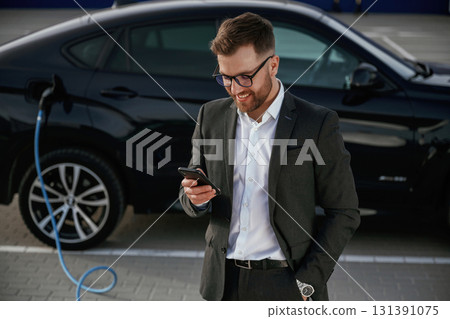 Smartphone in hand. Businessman in suit is near his black car outdoors Smartphone in hand. Businessman in suit is near his black car outdoors 131391075