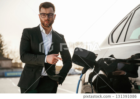 Pointing by finger to a socket with wire, charging. Businessman in suit is near his black car outdoors Pointing by finger to a socket with wire, charging. Businessman in suit is near his black car outdoors 131391086