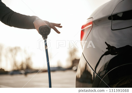 Blue wire for charging is in hand. Businessman in suit is near his black car outdoors Blue wire for charging is in hand. Businessman in suit is near his black car outdoors 131391088